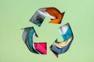 Recycle symbol cutout filled with colorful textile fabrics on a light green background, representing textile recycling.