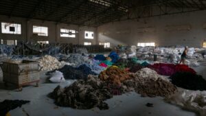 Interior of a large warehouse with piles of sorted and unsorted textile waste in various colors, highlighting the scale of post-consumer textile sorting operations.