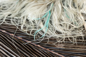 Close-up of off-white fibers with strands of turquoise and black-and-white patterned fibers resting on a pine-like textured brown surface.